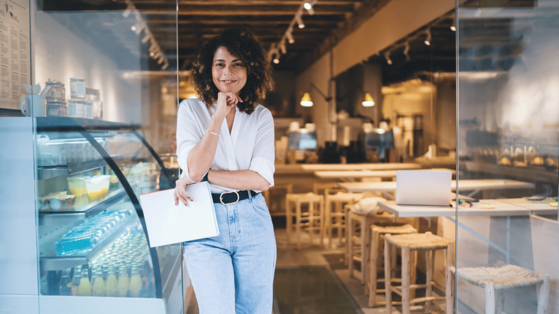 Woman standing confidently in a café, holding a clipboard, with display cases of beverages and pastries in the background, illustrating entrepreneurship and business management in the context of franchising.